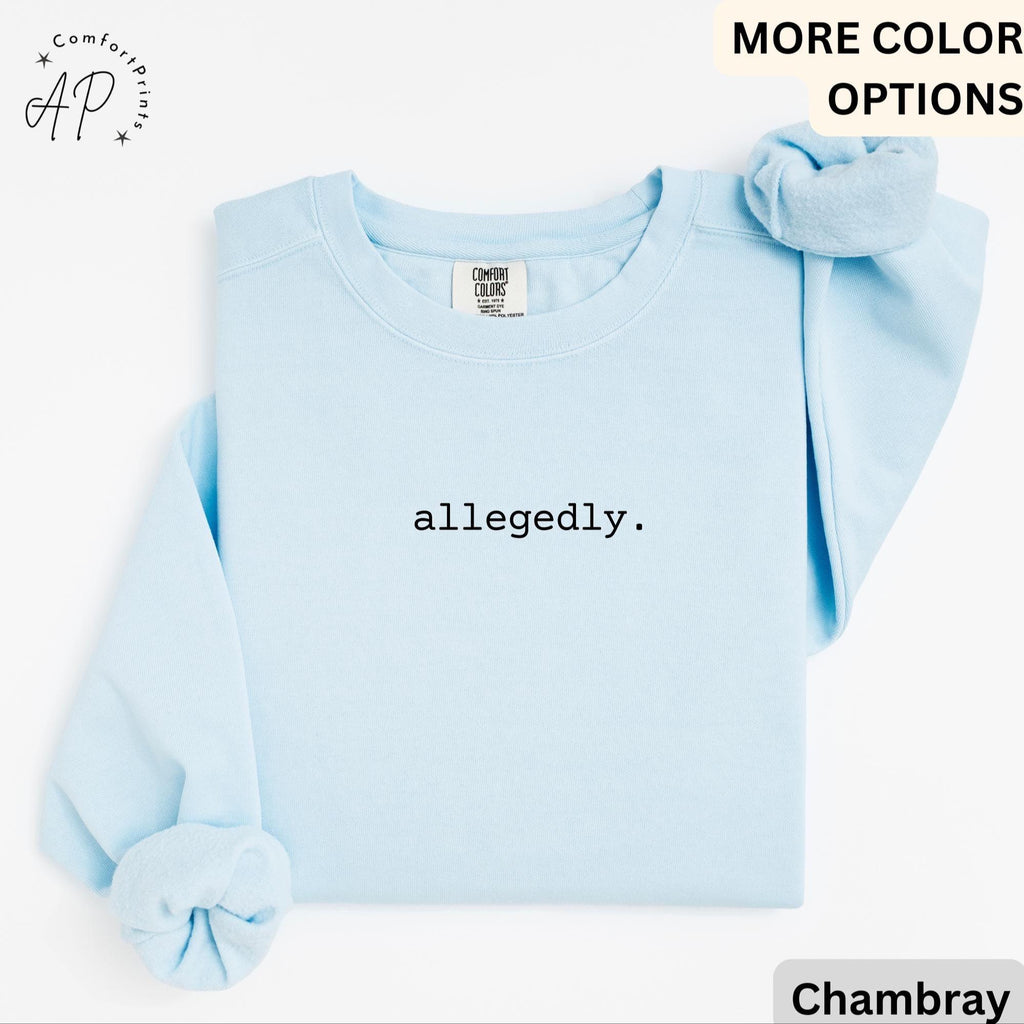 May include: A light blue sweatshirt with the word "allegedly" printed in black. The sweatshirt has long sleeves and a crew neck. A matching scrunchie is placed on the top right. The Comfort Colors brand tag is visible.