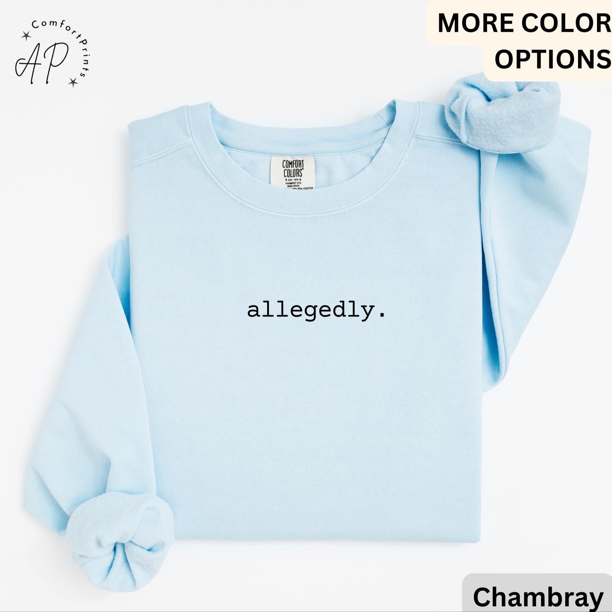 May include: A light blue sweatshirt with the word "allegedly" printed in black. The sweatshirt has long sleeves and a crew neck. A matching scrunchie is placed on the top right. The Comfort Colors brand tag is visible.