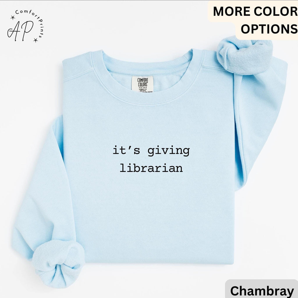 May include: Light blue sweatshirt with the text "it's giving librarian" in black. The sweatshirt has long sleeves and a crew neck. A matching scrunchie is placed on the top right. The brand logo "AP" is in the top left corner.