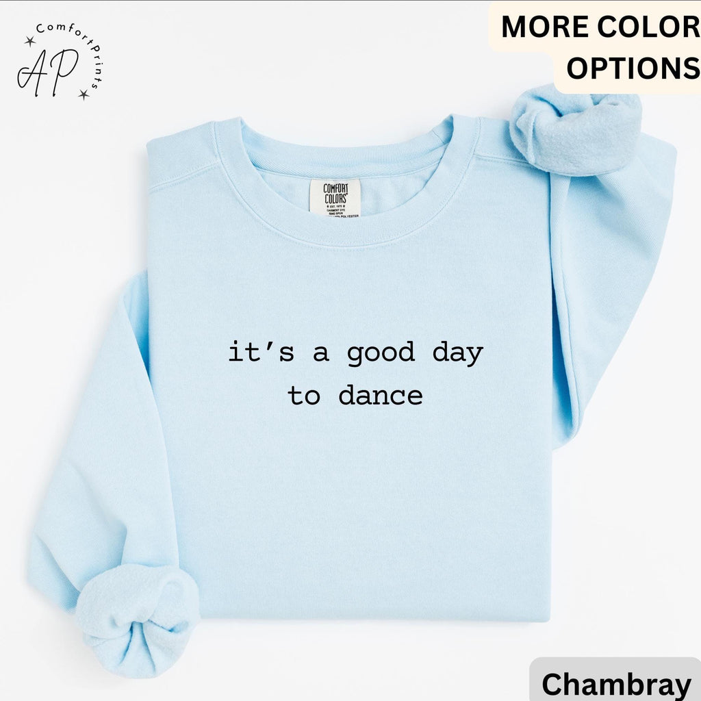 May include: Light blue sweatshirt with the text "it's a good day to dance" in black. The sweatshirt has long sleeves and a crew neck. A matching scrunchie is placed on the shoulder. The sweatshirt is made of a soft material.