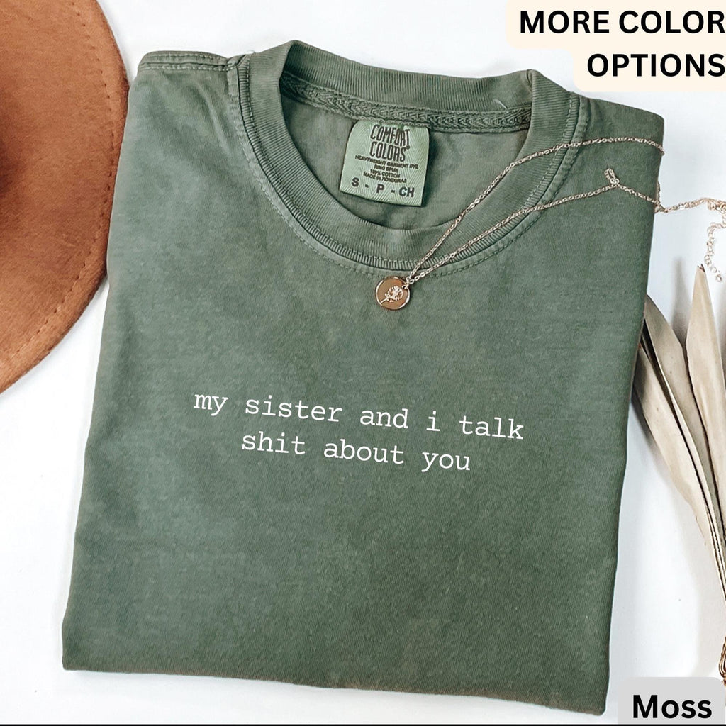 May include: A moss green t-shirt with the text "my sister and i talk shit about you" in white. A gold necklace with a small pendant rests on the shirt. The shirt is folded and has a Comfort Colors tag.