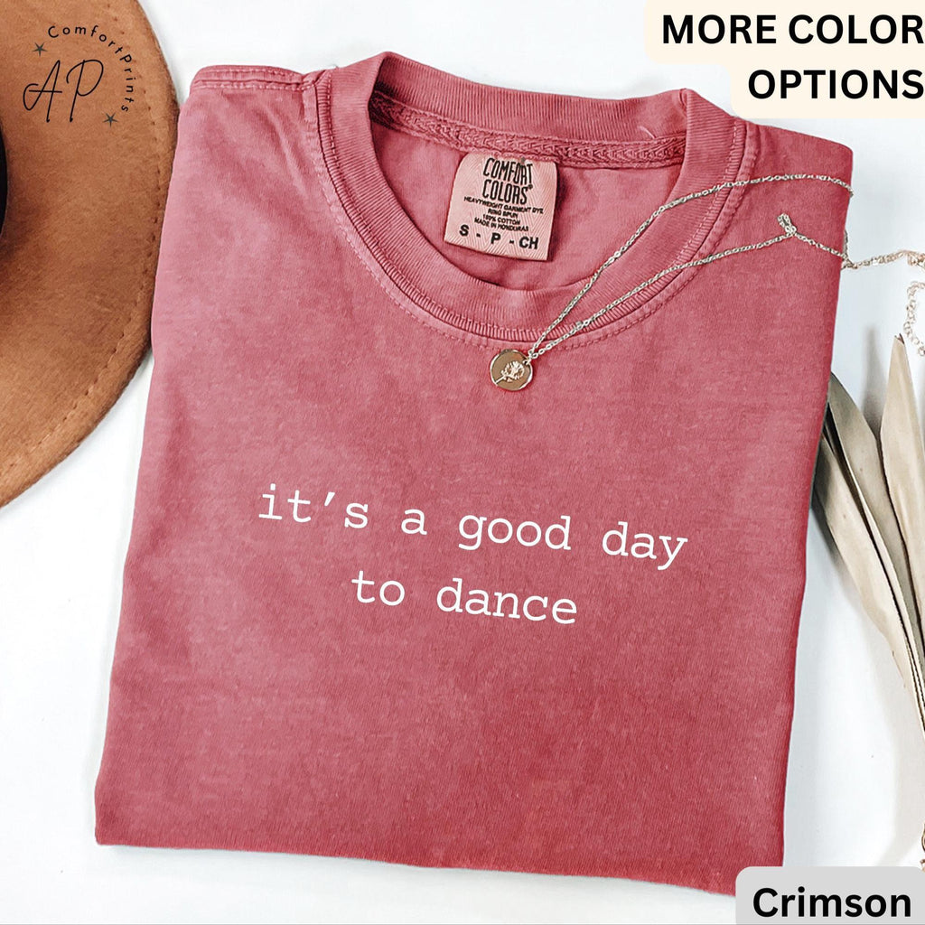 May include: Crimson-colored t-shirt with the text "it's a good day to dance" in white. The shirt has a crew neck and short sleeves. A silver necklace with a pendant is draped on the shirt.