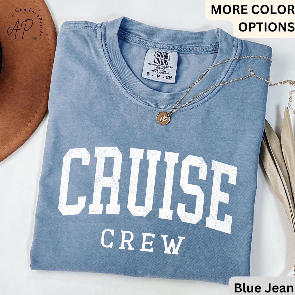 May include: A blue jean colored t-shirt with the words "CRUISE CREW" printed in white block letters. The shirt has a crew neck and short sleeves. A gold necklace with a pendant is draped across the shirt.