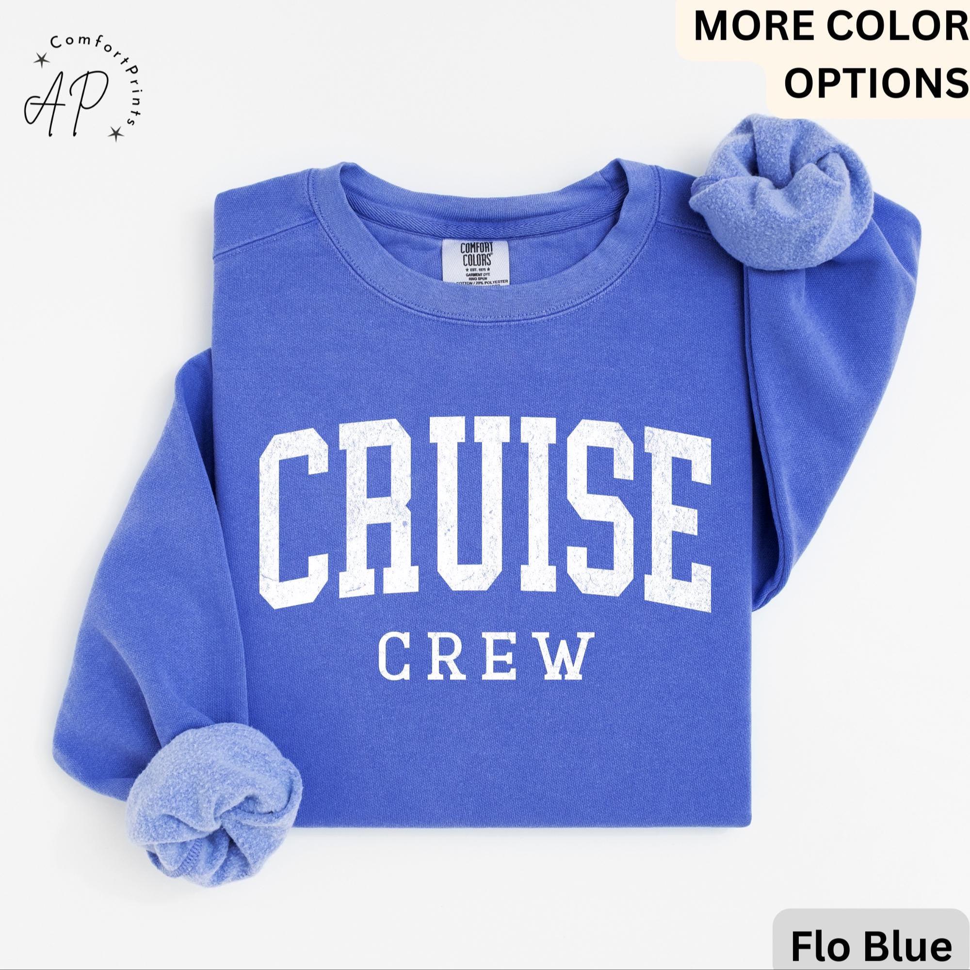 May include: Flo Blue sweatshirt with the words "CRUISE CREW" in white block letters. The sweatshirt is folded, with the sleeves cuffed. The Comfort Colors label is visible. The words "MORE COLOR OPTIONS" are in the top right corner.