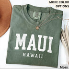 May include: A moss green t-shirt with the word "MAUI" printed in large white letters, and "HAWAII" below. A gold necklace with a pendant rests on the shirt. The shirt is folded, and the tag reads "Comfort Colors".