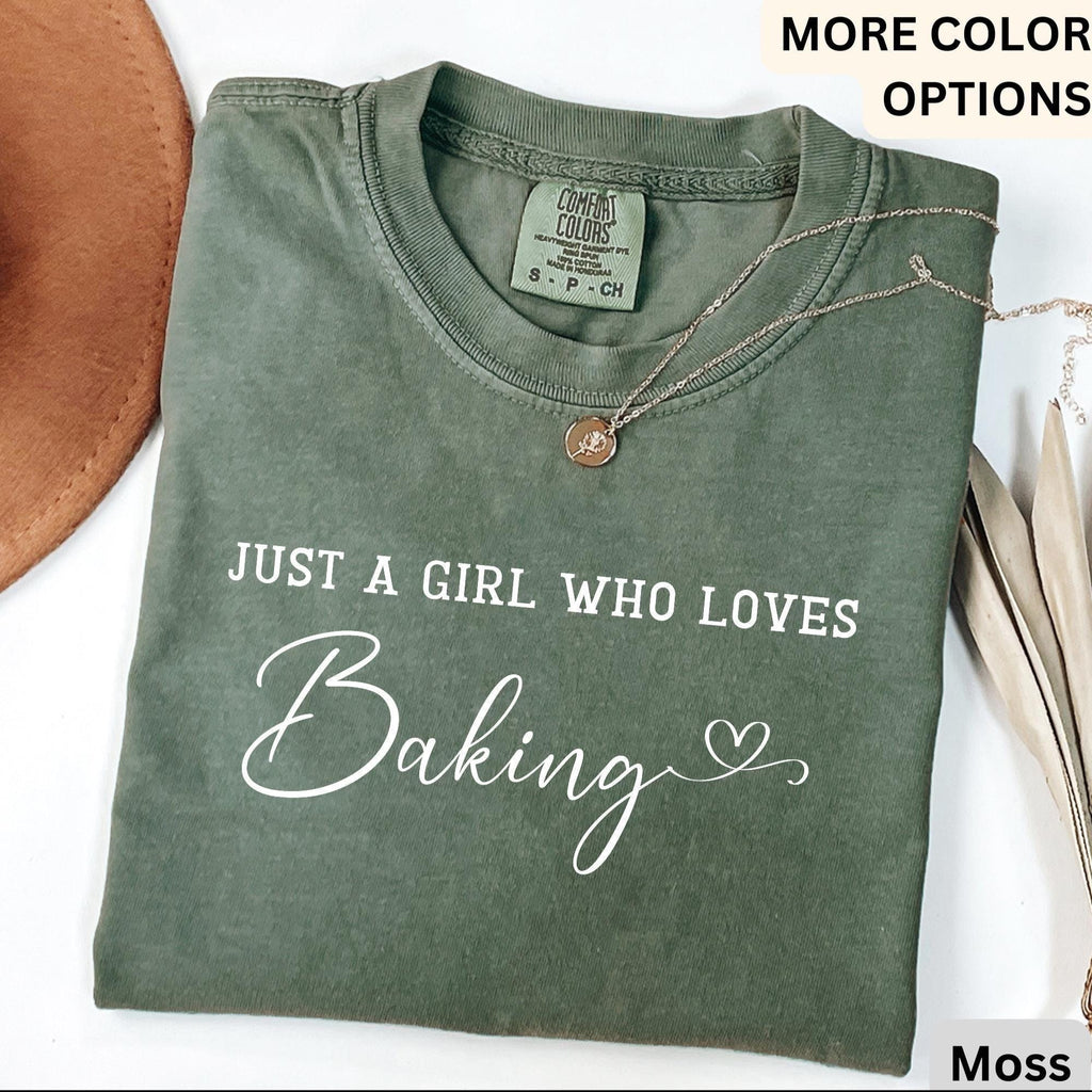 May include: Moss green Comfort Colors t-shirt with the text "JUST A GIRL WHO LOVES Baking" in white script. A gold necklace with a small pendant rests on the shirt. The shirt is folded and displayed with a brown hat.