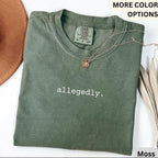 May include: Moss green t-shirt with the word "allegedly" printed in white. The shirt has a crew neck and short sleeves. A gold necklace with a small pendant is draped across the shirt. The shirt is folded and displayed with a brown hat.