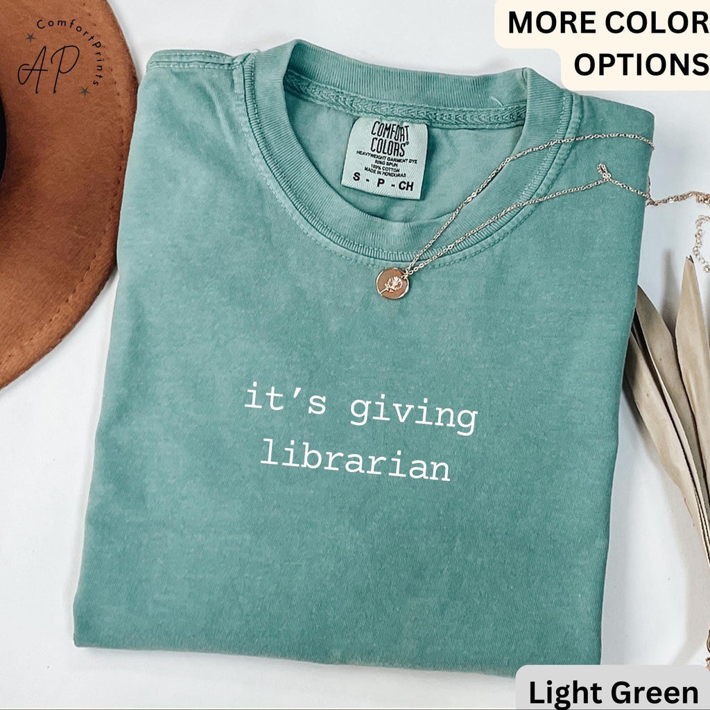 May include: Light green t-shirt with the text "it's giving librarian" in white. The shirt has a crew neck and short sleeves. A gold necklace with a pendant is draped across the shirt.