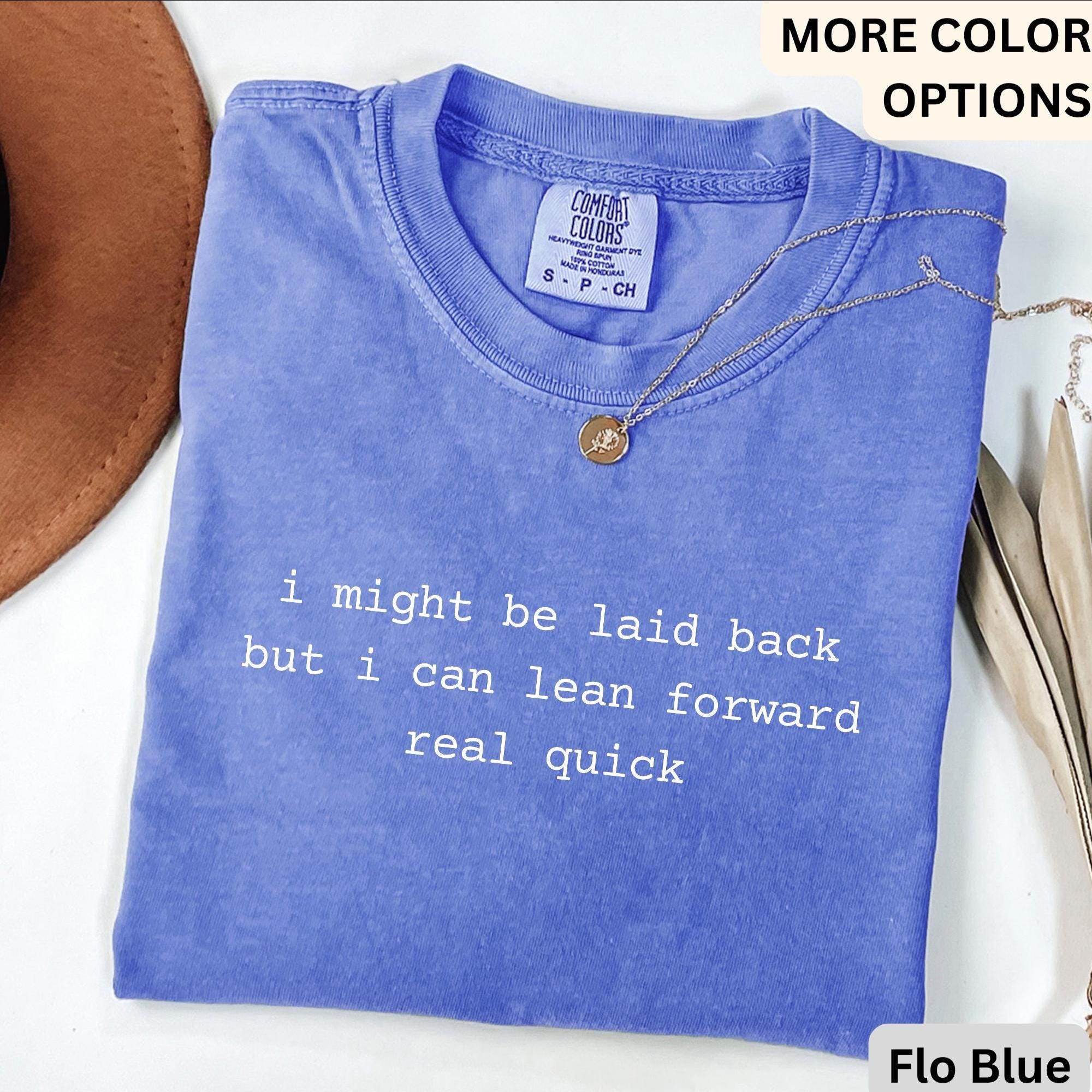 May include: A blue t-shirt with the text "i might be laid back but i can lean forward real quick" in white. The shirt has a crew neck and short sleeves. A gold necklace with a pendant is on the shirt.