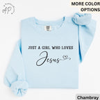 May include: Light blue sweatshirt with the text "JUST A GIRL WHO LOVES Jesus" in black script. The sweatshirt has long sleeves and a crew neck. A scrunchie in a matching light blue color is on top of the sweatshirt.