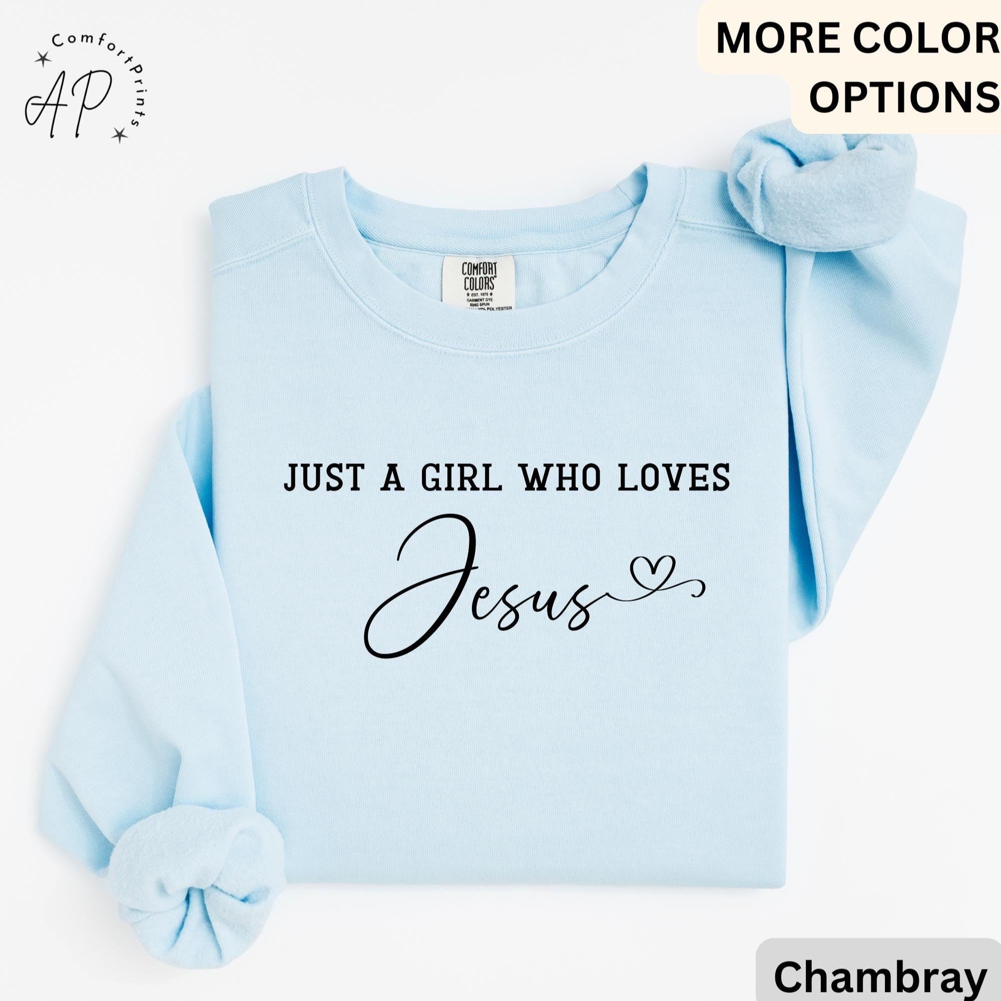 May include: Light blue sweatshirt with the text "JUST A GIRL WHO LOVES Jesus" in black script. The sweatshirt has long sleeves and a crew neck. A scrunchie in a matching light blue color is on top of the sweatshirt.