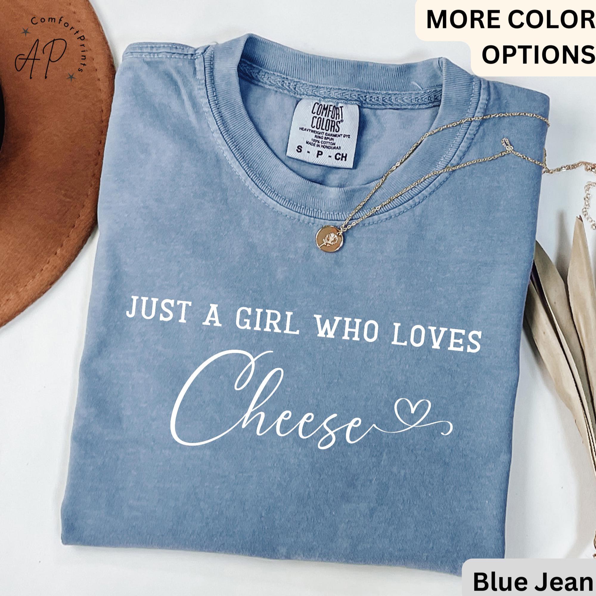 May include: Blue jean colored t-shirt with the text "JUST A GIRL WHO LOVES Cheese" in white script. The shirt has a crew neck and short sleeves. A gold necklace with a pendant is draped across the shirt.