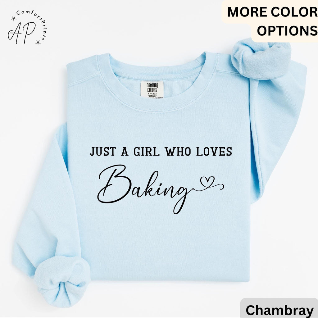 May include: Light blue sweatshirt with the text "JUST A GIRL WHO LOVES Baking" in black script. A matching light blue scrunchie rests on the shoulder. The sweatshirt is a Comfort Colors brand.