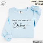 May include: Light blue sweatshirt with the text "JUST A GIRL WHO LOVES Baking" in black script. A matching light blue scrunchie rests on the shoulder. The sweatshirt is a Comfort Colors brand.