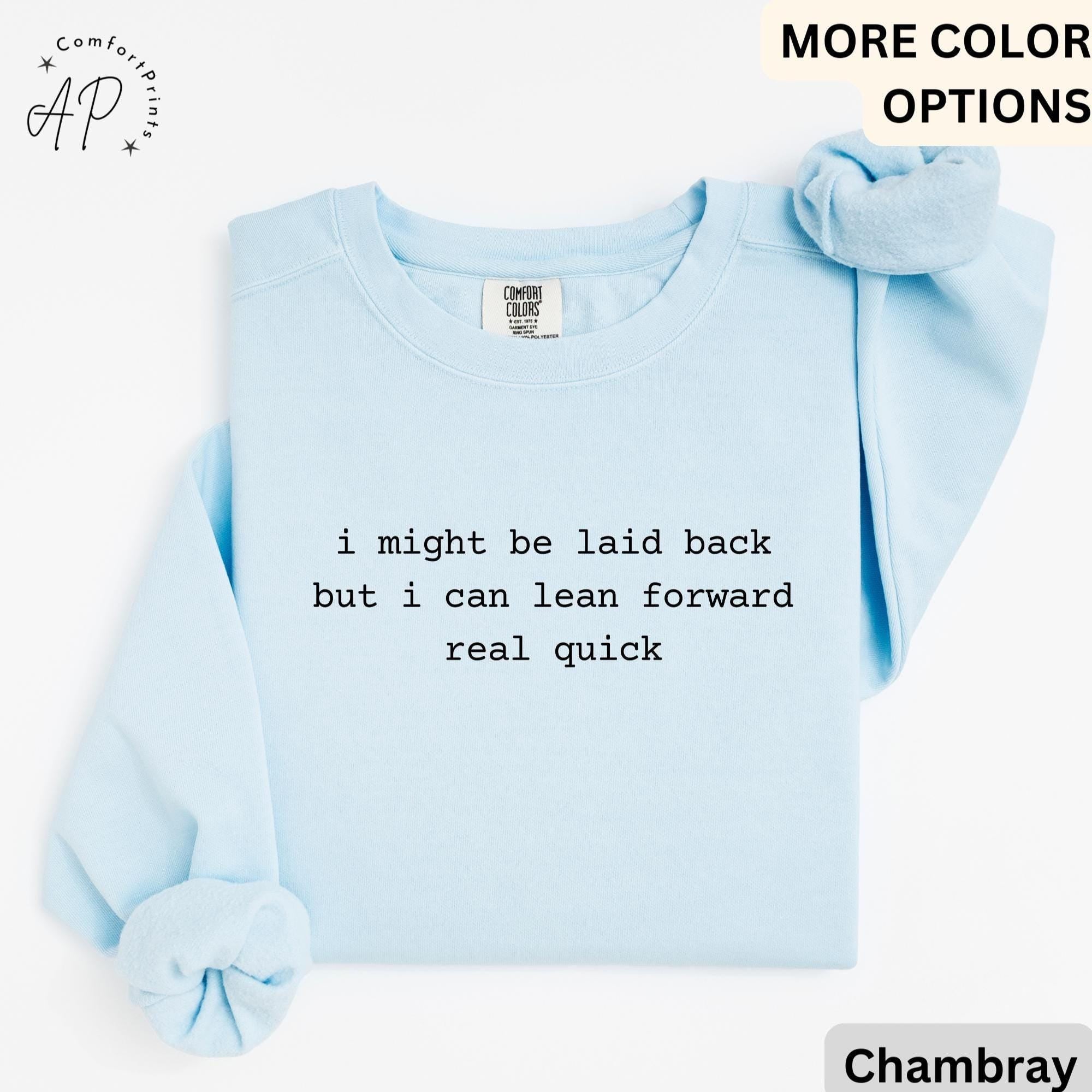 May include: A light blue sweatshirt with the text "i might be laid back but i can lean forward real quick." The sweatshirt has long sleeves and a crew neck. A scrunchie in a matching color is placed on the top right.