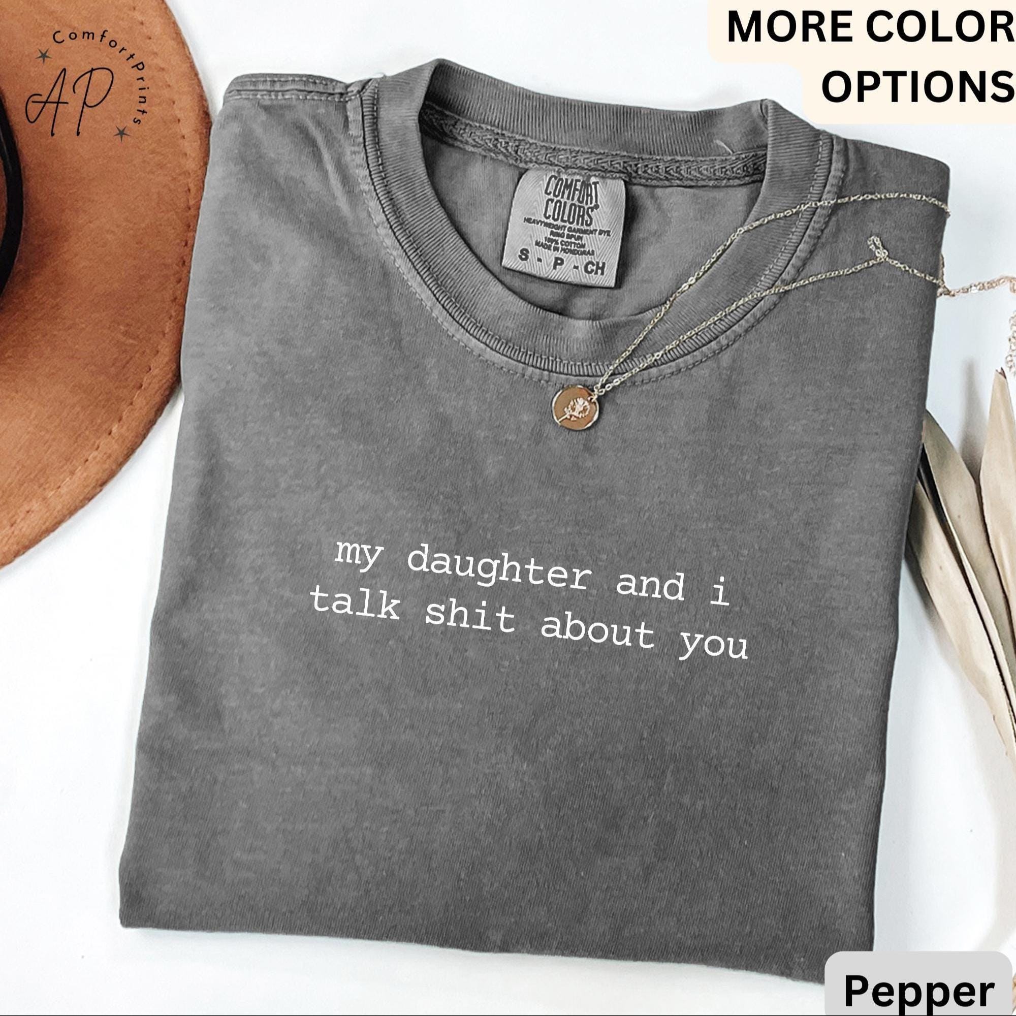 May include: Pepper-colored t-shirt with the text "my daughter and I talk shit about you" in white. The shirt has a crew neck and short sleeves. A silver necklace with a round pendant is on the shirt.